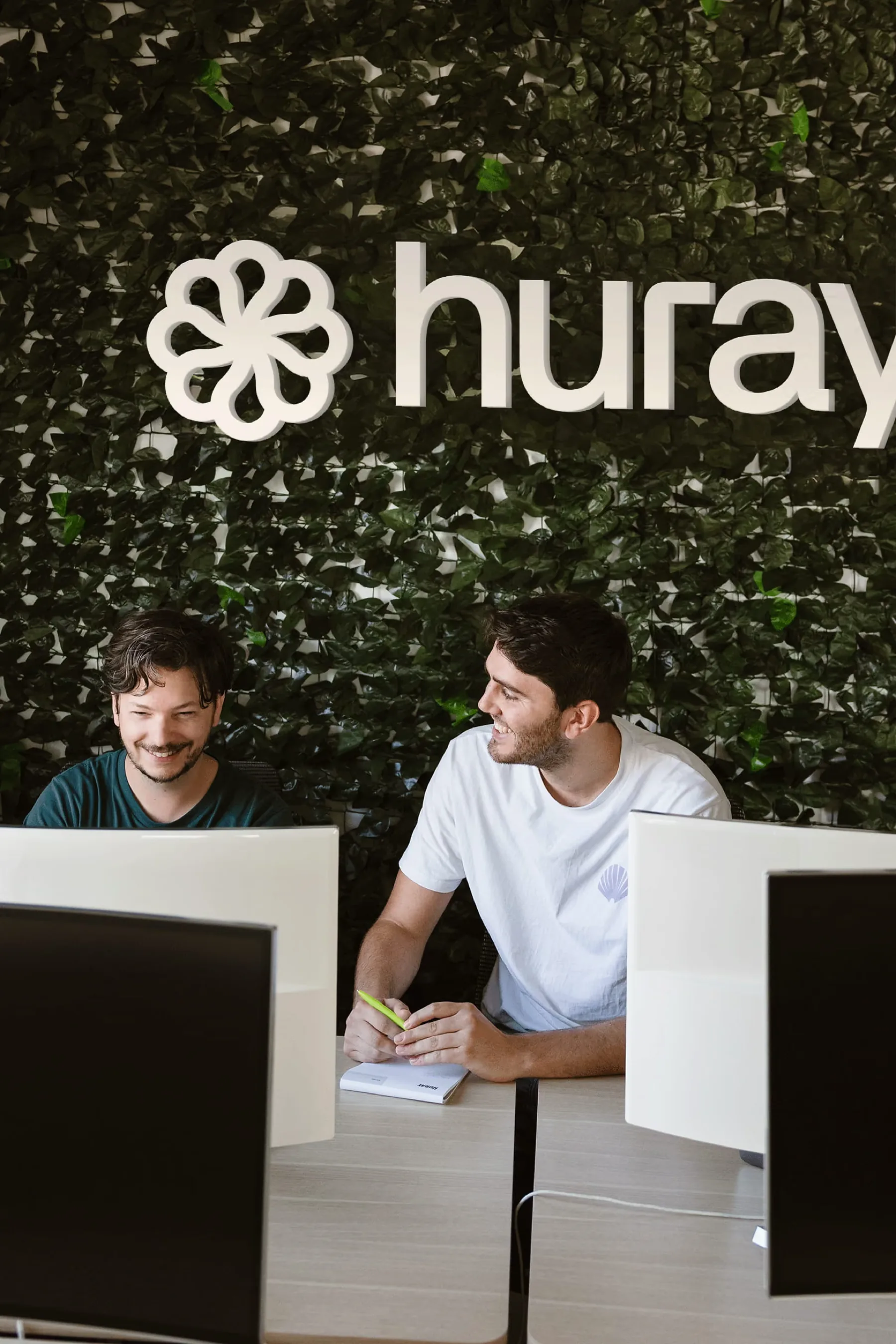 Twee collega's overleggen lachend achter hun schermen voor de groene merkwand van Huray.